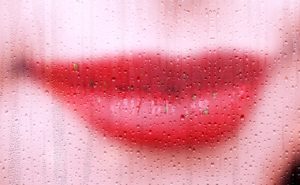 bright red lips in a slight smirk behind a window covered in droplets of water