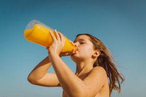 A young child with no shirt and light brown hair against a blue sky is swallowing gulps of orange juice from a clear plastic bottle. 